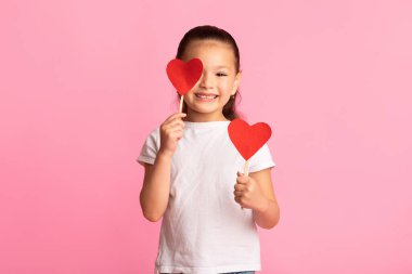 Çubuklarda iki kırmızı kalple oynayan sevimli Japon kız bir gözünü kapatıp kameraya bakıyor, pembe duvar.