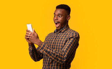 Mobil Teklif. Zeki telefon ekranına heyecanla bakan komik siyah adam, çevrimiçi haberlere duygusal tepki veren, ağzı açık duran, sarı arka planda izole edilmiş, boşluğu kopyalayan.