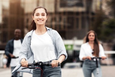 Bir grup genç kalabalık bir şehir caddesinde scooterla geziyor, açık hava atmosferinin tadını çıkarıyorlar. Gülüşleri, şehir manzarasında gezinirken heyecanı ve özgürlüğü yansıtıyor..