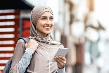 Türbanlı olumlu Arap kız kulaklık ve en yeni akıllı telefonu kullanıyor, sokakta yürüyor ve müzik dinliyor, boş alan arıyor.
