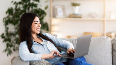 Favori eğlencesi. Neşeli Asyalı kadın dizüstü bilgisayarla koltukta rahatlıyor, internete bakıyor ya da film seyrediyor, hafta sonunun keyfini çıkarıyor.