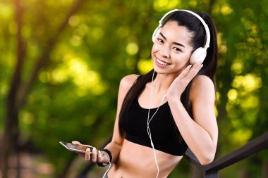 Asyalı bayan koşucu eğitimde ara veriyor, dışarıda akıllı telefon ve kulaklıkla müzik dinliyor, boşluğu kopyalıyor.