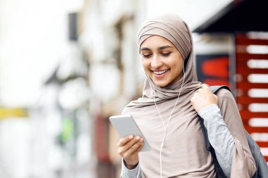 Her zaman bağlı kal. Türbanlı güzel bir kız kulaklık ve en yeni akıllı telefonu kullanıyor, sokakta yürüyor, boş bir alanda.
