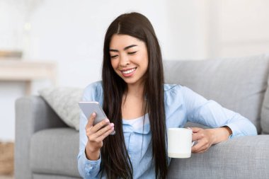 Olumlu Koreli kız akıllı telefondan mesaj atıyor ve evde sabah kahvesinin tadını çıkarıyor, oturma odasında yerde oturuyor, boş bir yerde oturuyor.
