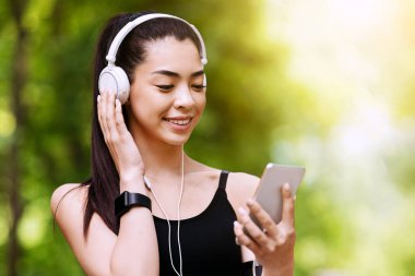 Online Müzik Uygulaması. Akıllı telefonu ve kulaklığı olan neşeli koşucu kız. Açık havada koşmak için çalma listesi seçiyor.