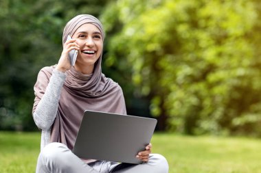 Parkta çalışan, dizüstü bilgisayar kullanan ve müşterilerle telefonda konuşan, serbest çalışan pozitif Arap kız.