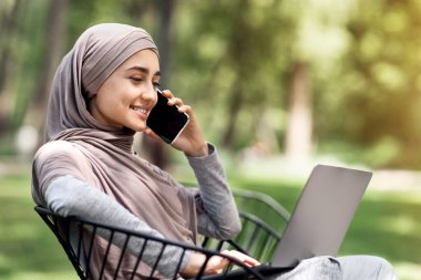 Tesettürlü neşeli Arap kız telefonda konuşuyor ve parkta bankta otururken dizüstü bilgisayar kullanıyor.