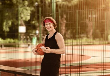 Açık hava basketbol sahasında cep telefonu ve topu olan mutlu bir milenyum atleti.