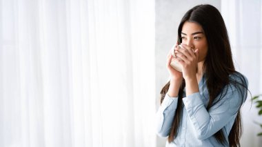 Rahatlamış Asyalı kadın çay içiyor ve evden manzaranın tadını çıkarıyor, sabah kahvesini içiyor, sıcak bardakla ellerini ısıtıyor, rahat oturma odasında dinleniyor, izinli bir gün geçiriyor, uzay kopyası yapıyor.