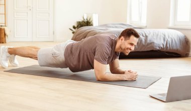 Formda Adam Evde Çevrimiçi Antrenman sırasında dizüstü bilgisayar egzersizi yapıyor. İçerde egzersiz yapan bir adam var. Spor yaşam tarzı, erkek spor rutini ve kilo kaybı konsepti.