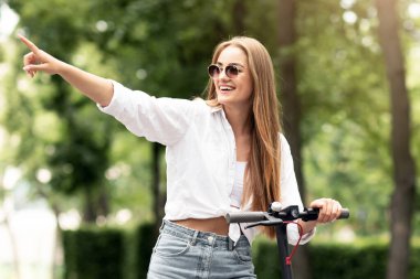 Arkadaşlık kavramı. Gülümseyen genç kız e-scooterıyla dikiliyor ve arkadaşlarına manzara gösteriyor.
