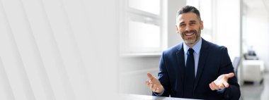 Takım elbiseli orta yaşlı bir işadamı iyi aydınlatılmış bir ofiste bir tartışma yapıyor. Kendinden emin ve cana yakın görünüyor, çağdaş bir ortamda başarılı ve profesyonel bir yaşam tarzı sergiliyor..