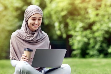 Güzel Arap kız halka açık parkta dizüstü bilgisayar kullanıyor, çimlerin üzerinde oturuyor, kahve içiyor, boş bir alanda.