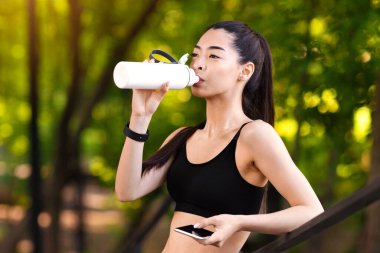 Parkta koştuktan sonra su içen mutlu genç Asyalı kız, akıllı telefonu tutuyor ve korkuluklara yaslanıyor, dışarıda egzersiz yapıyor, boş alan arıyor.