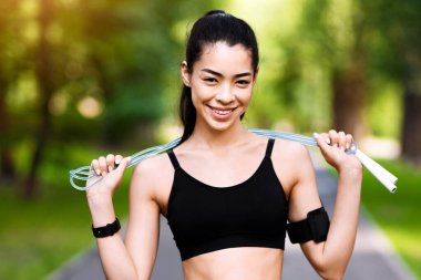 Açık havada kardiyo eğitimi. Pozitif Asyalı kız ip atlıyor, parkta egzersiz yapmaya hazır, kameraya bakıyor ve gülümsüyor,