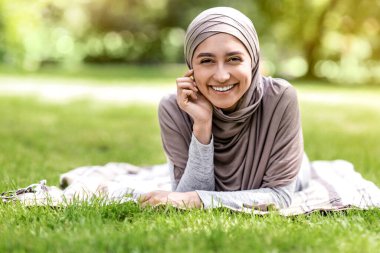 Tesettürlü neşeli Arap kız parkta çimlere uzanıyor, kameraya gülümsüyor.