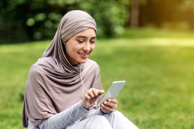 Kulaklık takan, cep telefonu kullanan, parkta oturan mutlu Arap kadın.