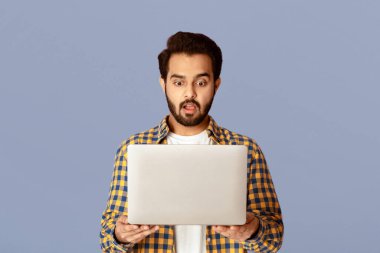 Leylak arka planında ağzı açık bir şekilde dizüstü bilgisayar ekranına bakan şok olmuş Hintli adam.