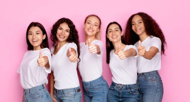 Çok kültürlü beş kadından oluşan bir grup pembe arka planda birlikte poz vererek el hareketlerini gösteriyor. Çeşitli bayanların başparmaklarıyla bir şeyi onaylayan stüdyo fotoğrafları. Panorama