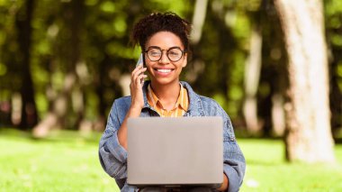 Her zaman temastayız. Cep telefonuyla konuşan siyah bir kadın internette bilgisayarda çalışıyor parkta oturuyor, kameraya bakıyor. Serbest Yaşam Tarzı, Uzak İnternet Çalışma Konsepti. Panorama