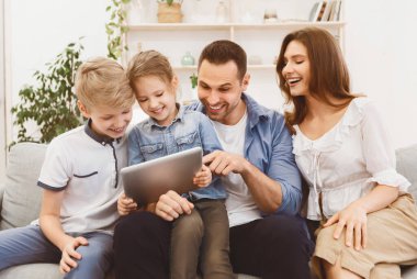 Ebeveynler ve çocuklar dijital tablet kullanıyorlar. Evdeki koltukta birlikte oturuyorlar. Aile Eğlencesi Konsepti