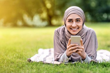 Akıllı telefonunu kullanarak gülen Müslüman kadın, komik içerikler okuyor, parkta dinleniyor, boş bir alanda.