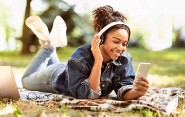 Neşeli Siyah Milenyum Kadını Cep Telefonu ve Kulaklık Kullanıyor Ekoseli Hafta Sonu Parkta Uzanırken Dinleyen Müzik Dinliyor. Sonbahar Şarkı Listesi Konseptinin keyfini çıkarın