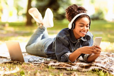 Neşeli siyahi kız dışarıda, parkta ekose yatarken dizüstü bilgisayarla telefon kullanmayı öğreniyor. Öğrenciler Yaşam Tarzı Konsepti