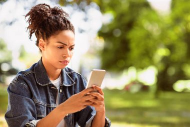 Haberler kötü. Açık havada oturan hoşnutsuz Afro-Amerikan Kadın Akıllı Telefon kullanarak Olumsuz Mesaj Okuyor. Cep telefonu sorunu ve sorunları kavramı. Metin için Boşluk