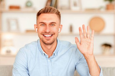 Selamlama Kavramı. Gülümseyen yakışıklı adamın portresi. El sallayarak merhaba diyor. Kameraya bakıyor, evde oturuyordu, kablosuz kulaklık takıyordu. Adam merhaba diyor, gülüyor, geçmişi bulanık.