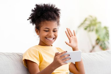 Akıllı telefonlu Afrikalı Amerikalı genç kız evdeki koltukta oturmuş el sallayarak selam veriyor. Uzak İletişim Konsepti