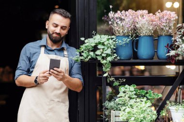 Küçük işletme sahibi için sosyal medya, reklam ve online teklif. Gülümseyen sakallı bin yıllık Avrupalı erkek önlüklü akıllı telefonuyla çiçekçi dükkanının ön kapısında farklı bitkiler ve boş alan olan akıllı telefonu izliyor.