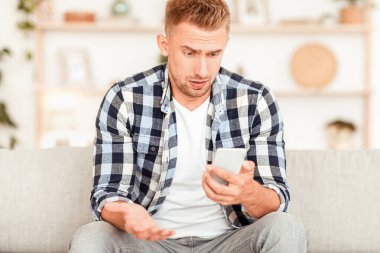 Kötü Haber Konsepti. Akıllı telefona bakan şaşırmış bir adamın ön portresi, sosyal medyadan veya mesajdan hayal kırıklığına uğramış, evdeki kanepede oturan. Cep telefonu kullanan kafası karışmış bir adam.