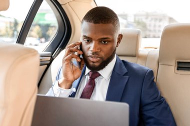 Zengin afro-amerikan işadamı şık takım elbiseli, arabada çalışıyor, dizüstü bilgisayar kullanıyor ve akıllı telefonla konuşuyor, internette çalışıyor, kopyalama alanında çalışıyor. Yoğunlaştırılmış siyah yöneticinin telefonu var.