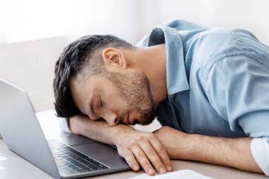 İşte uyu. Modern ofisin işyerinde çok çalışan yorgun bir adam laptopla ve raporlarla çalıştıktan sonra uyukluyor, masa başında oturuyor, baş aşağı oturuyor, iş yorgunluğu çekiyor.