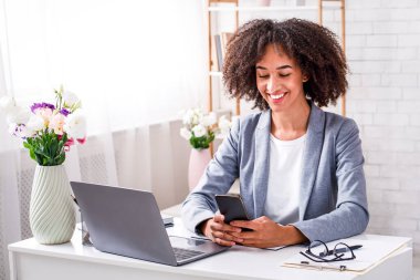 Coronavirus karantinası sırasında evde yeni iş ve moda blogu. Akıllı telefondan yazı yazan mutlu Afro-Amerikalı kadın oturma odasında dizüstü bilgisayarla oturuyor ve tezgahta giysileri var.