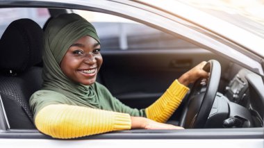 Araba sigortası konsepti. Gülümseyen Afrikalı Müslüman kadın modern araçta oturuyor, yeni arabasını sürmekten zevk alıyor, direksiyonu tutuyor, kameraya poz veriyor ve gülümsüyor açık pencereden, boş bir pencereden,