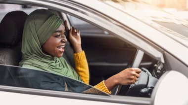 Sürücü eğlencesi. Neşeli siyah Müslüman kadın tesettürde müzik dinliyor ve arabada şarkı söylüyor, heyecanlı Afrikalı İslamcı kadın şehirde sürmekten zevk alıyor, direksiyonu tutuyor ve yola bakıyor, yandan bakıyor.