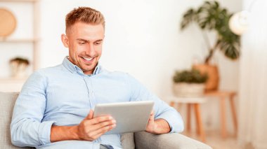 Boş zaman kavramı. Evdeki kanepede otururken dijital tablet kullanan tatmin olmuş bir adamın portresi, bulanık arka plan. Gülümseyen adam internetten alışveriş yapıyor, internette geziniyor, kanepede dinleniyor.