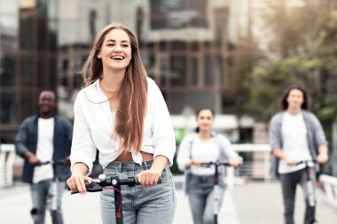 Gençlik Konsepti. Genç ve güzel bir kız yazın elektrikli scooter kullanıyor, arkadaşlarıyla eğleniyor.