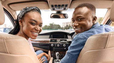 Yeni arabalarında poz veren, arka koltuğa bakıp test sürüşü yaparken kameraya gülümseyen mutlu bir Afro-Amerikan çifti. Neşeli siyah adam ve kadın yaz tatiline arabayla gidiyorlar.
