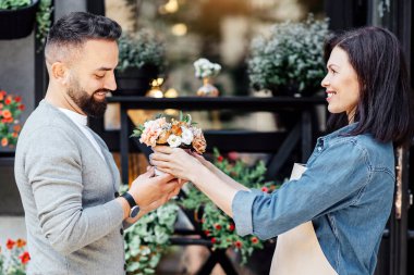 Çiçek butiği, çiçekçilik, küçük işletme ve dekorasyon konularında yaratıcı bir uğraş. Önlüklü genç bayan satıcı saksıda bitkileri olan çiçekçi dükkanının yakınındaki müşteriye buket veriyor.