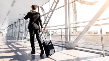 Businessman in black suit walking by airport with suitcase, back view, sun flare, panorama with copy space. Unrecognizable man having business trip, talking with business partners on mobile phone