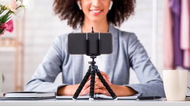 Covid-19 salgını sırasında takipçiler için giyim ve yayınlama videosu. İş elbiseli, kablosuz kulaklıklı, oturma odasındaki akıllı telefon kamerasına bakan dost canlısı Afro-Amerikan kadın.