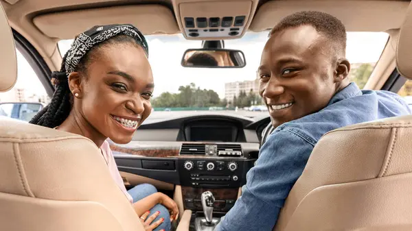 Yeni arabalarında poz veren, arka koltuğa bakıp test sürüşü yaparken kameraya gülümseyen mutlu bir Afro-Amerikan çifti. Neşeli siyah adam ve kadın yaz tatiline arabayla gidiyorlar.
