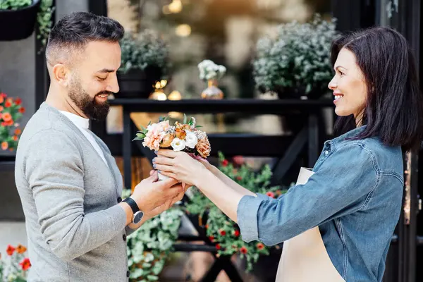 Çiçek butiği, çiçekçilik, küçük işletme ve dekorasyon konularında yaratıcı bir uğraş. Önlüklü genç bayan satıcı saksıda bitkileri olan çiçekçi dükkanının yakınındaki müşteriye buket veriyor.