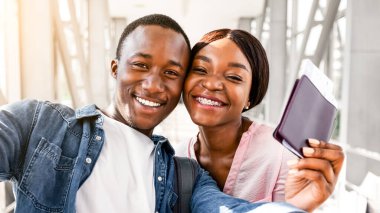 Havalimanı terminalinde selfie çeken mutlu siyah bir çiftin portresi, Afrika kökenli Amerikalı aşıkların ellerinde pasaport ve bilet tutmaları, uçuşlarını beklerken kameraya gülümsemeleri,