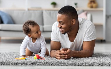 Genç siyah baba ve sevimli bebeği evde birlikte rahatlarken, Afrikalı Amerikalı baba ve tatlı yeni doğan oğlu oturma odasında halıya uzanmış, bebek çocuk babaya merakla bakıyor.
