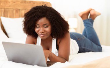 Çevrimiçi sohbet. Gülümseyen Afro-Amerikan kadın evinde yatağında dizüstü bilgisayarını kullanıyor. Mutlu pozitif siyah kadın video izliyor, internete ve sosyal medyaya bakıyor ya da uzaktan çalışıyor.