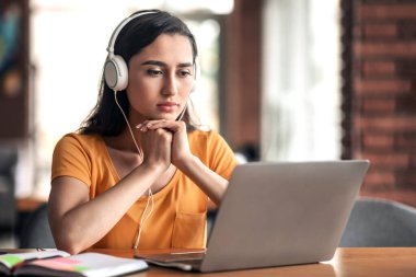 Yoğunlaşmış genç Arap kadın çevrimiçi eğitim dersi veriyor, dizüstü bilgisayar, kulaklık, kafenin içi, boş alan kullanıyor. İnternette okuyan ciddi bir kız öğrenci kafeteryada dizüstü bilgisayarın önünde oturuyor.